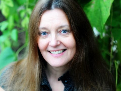 Gerda stevenson in nature smiling. She has long brown hair and blue eyes.