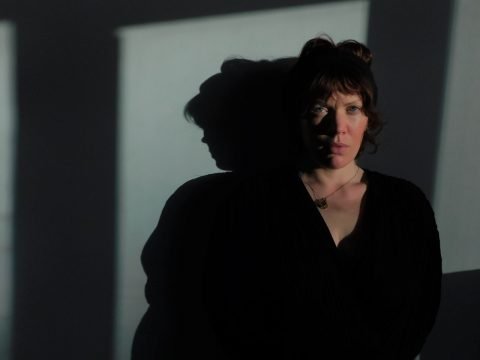 A headshot of poet Rachel McCrum (a woman with short dark hair). She is standing in shadow and wearing a black shirt. Her shadow is visible behind her.