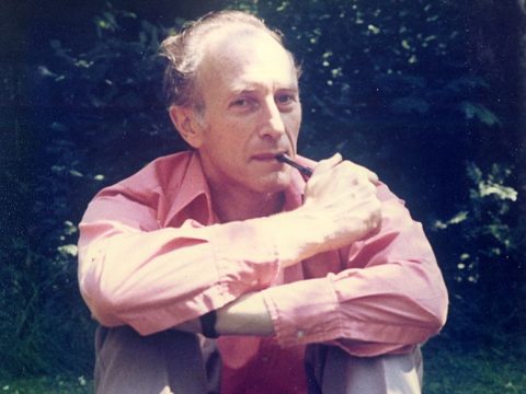 Norman Kreitman in his garden, smoking a pipe and wearing a red shirt.