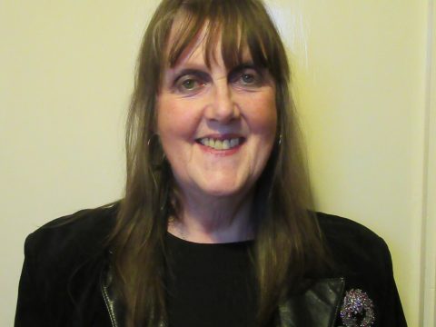 A headshot of Màiri NicGumaraid (Mary Montgomery). She has long blond hair and is wearing a black blazer with a brooch.