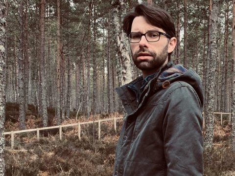 Dark haired man wearing glasses and a waxed jacked stands in front of trees in a wooded area