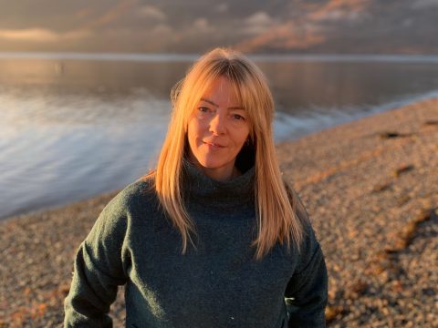 Woman with long blonde hair, standing in front of a loch on a sunny day