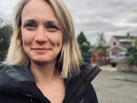 A smiling woman with blonde shoulder length hair, wearing a dark anorak and standing outside in a residential area during the day.