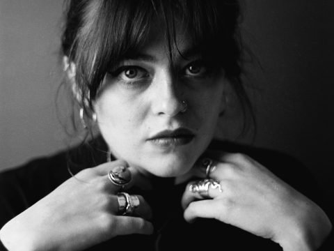 Young woman with dark hair tied back, and wearing a black high neck jumper. She is wearing several large rings and her hands are sitting under her chin.