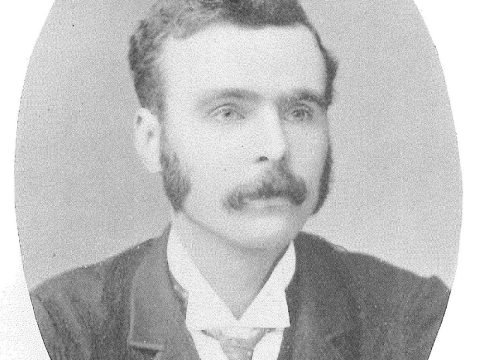 Man from the 19th century, with short dark hair, whiskers and moustache, posing for photograph wearing a dark jacket and waistcoat with a white shirt and polka dot patterned tie.