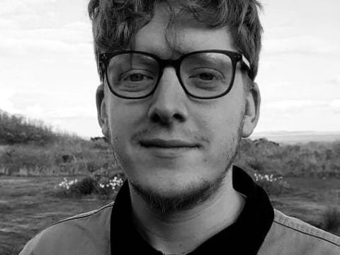 young man with curly hair, beard and dark rimmed glasses standing in front of a rural landscape