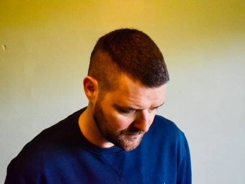 White man with short dark hair and beard, wearing a blue sweatshirt is standing in front of a light coloured wall