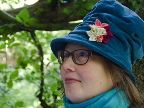 Young woman wearing a blue velvet hat with 3 pink and cream flowers, a blue scarf and glasses standing in front of a tree