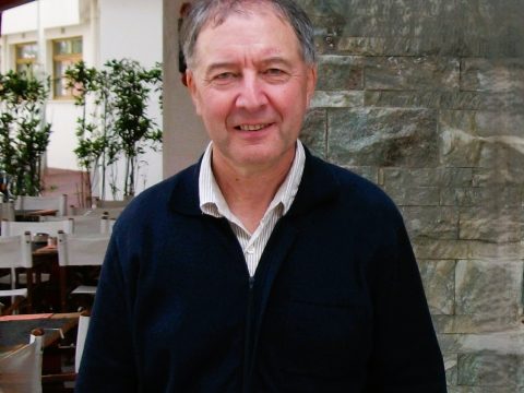 Older man wearing a navy jumper and light coloured shirt, standing in front of stone wall and cafe